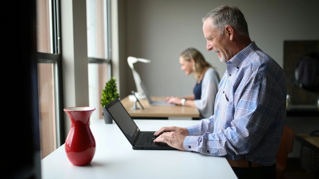 A Waltz member working on a laptop near a window while a teammate works in the background.